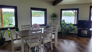 A dining area with a wooden table and white chairs, large windows with a view of a lake, and potted plants.