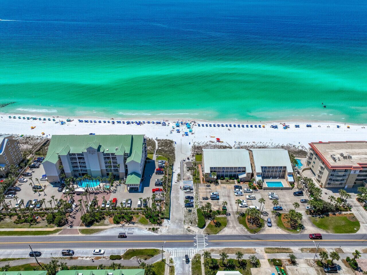 An aerial view of a coastal hotel with multiple buildings, a beach with umbrellas, and a parking lot.