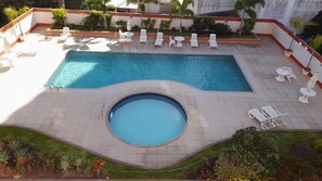 A rectangular swimming pool with a circular hot tub in the center, surrounded by white lounge chairs and small round tables. - Via Park Flat PARAÍSO (Campos)