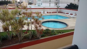 Piscina com duas áreas, rodeada por espreguiçadeiras brancas e palmeiras.