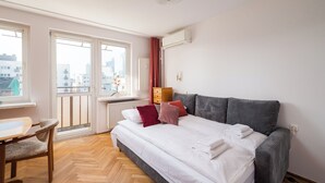 A hotel room with a bed, a wooden chair, a window with a view of buildings, and a radiator. - Złota Standard Studio (Warszawa)
