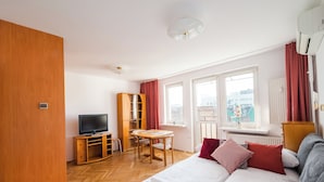 A hotel room with a bed, a TV, a wooden wardrobe, a desk with a chair, and large windows. - Złota Standard Studio (Warszawa)