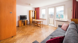A modern living room with a wooden floor, a grey sofa, a wooden TV stand, a dining table with chairs, and a large window with a view of buildings. - Złota Standard Studio (Warszawa)