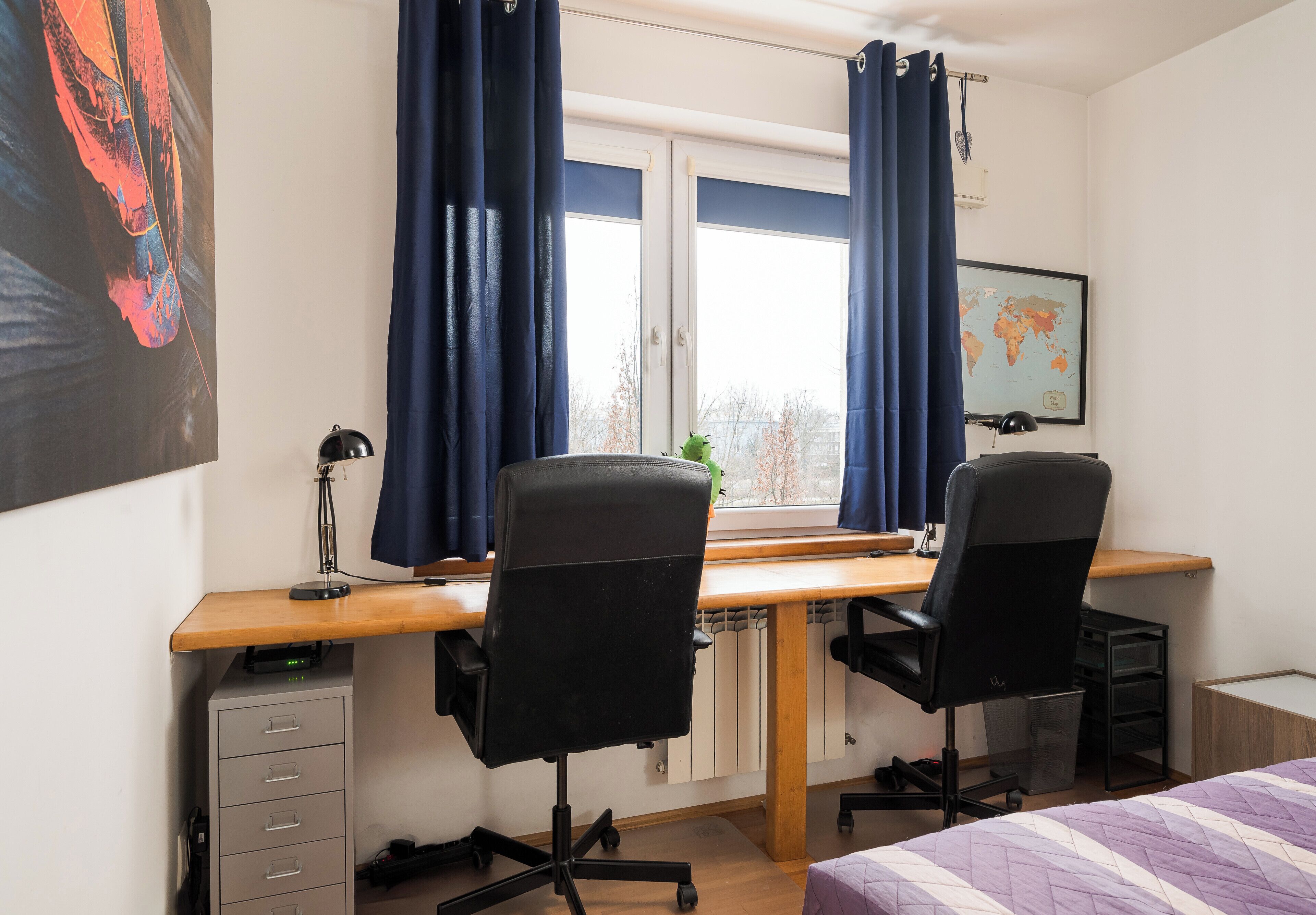 A bedroom with a bed, a desk with two chairs, a radiator, a window with curtains, a map on the wall, and a filing cabinet.