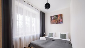 Ein Schlafzimmer mit einem Bett, weißen Kissen, einer grauen Decke, einer schwarzen Pendelleuchte, einem Gemälde an der Wand und transparenten Vorhängen.