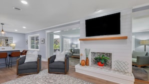 Uma sala de estar moderna com lareira, dois poltronas, uma TV sobre a lareira e uma cozinha visível através de uma porta aberta.