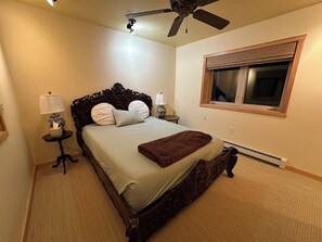 Une chambre à coucher avec un grand lit, deux tables de chevet, un ventilateur de plafond, une fenêtre avec des stores et un radiateur.