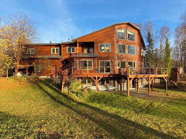 Une maison en bois à deux étages, dotée d’une terrasse et entourée d’arbres et d’herbe.