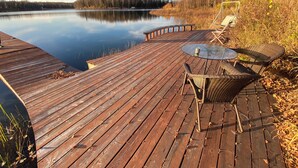Een houten terras met een glazen tafel en rieten stoelen met uitzicht op een rustig meer, omgeven door bomen en een bos.