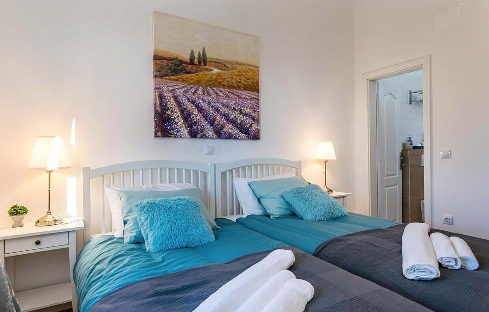 Habitación de hotel con dos camas, cada una con ropa de cama color turquesa y almohadas mullidas. En la pared, hay un cuadro de un campo, una mesita de noche con una lámpara y una puerta que da a otra habitación.