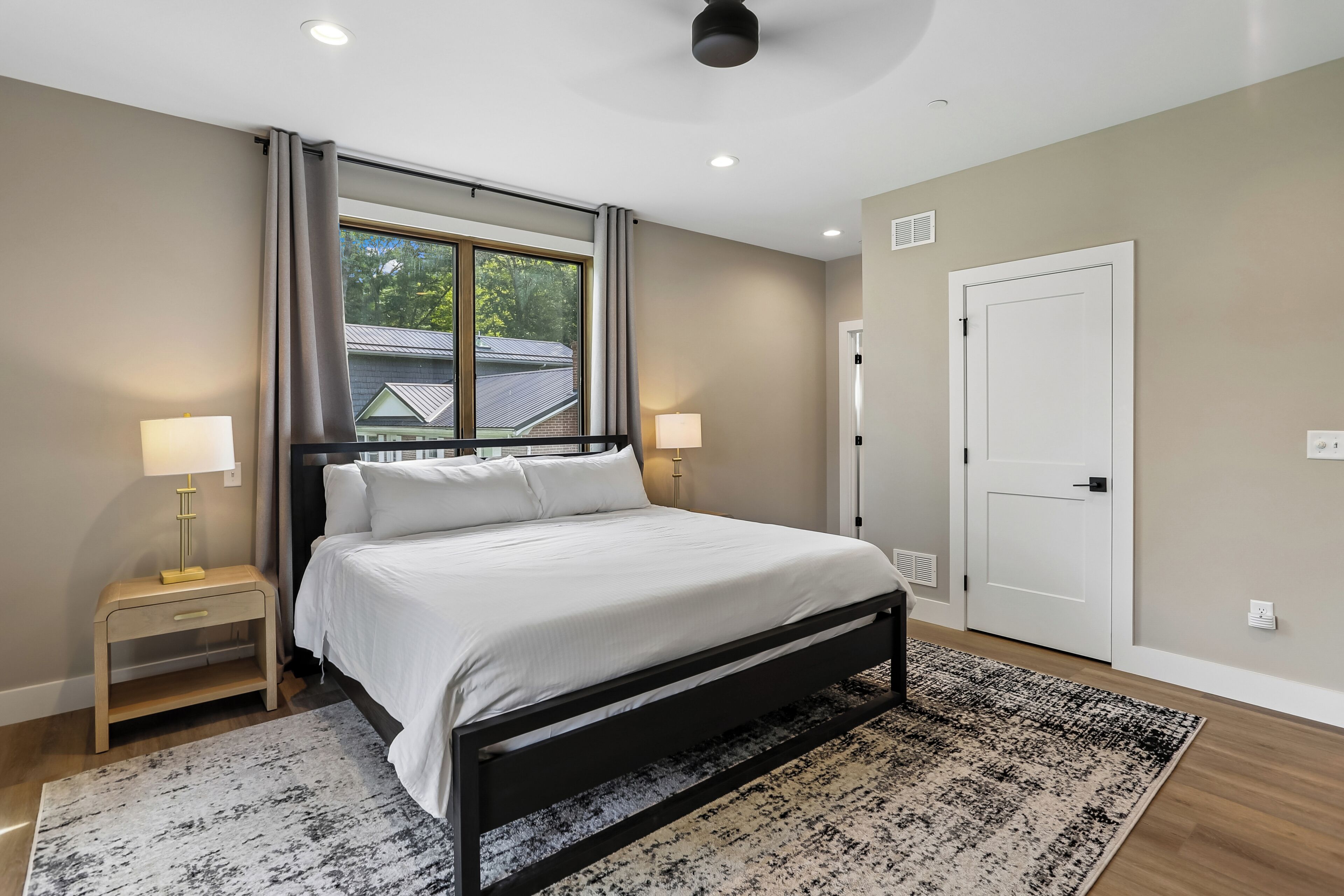 Une chambre à coucher avec un lit, des tables de chevet, une fenêtre avec des rideaux, une porte et un luminaire au plafond.