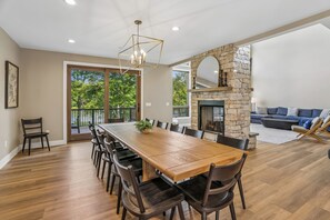 Une salle à manger spacieuse avec une longue table et des chaises en bois, une cheminée en pierre et une vue sur l’extérieur grâce à de grandes fenêtres.