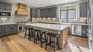 A modern kitchen with grey cabinets, a large island with stools, stainless steel appliances, and a window with blinds. - New Construction! 7 bedroom luxury lakefront  home in golf neighborhood! (Swanton)