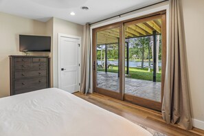 A bedroom with a bed, a dresser, a television, and a sliding glass door leading to a patio with a view of a lake and trees. - New Construction! 7 bedroom luxury lakefront  home in golf neighborhood! (Swanton)