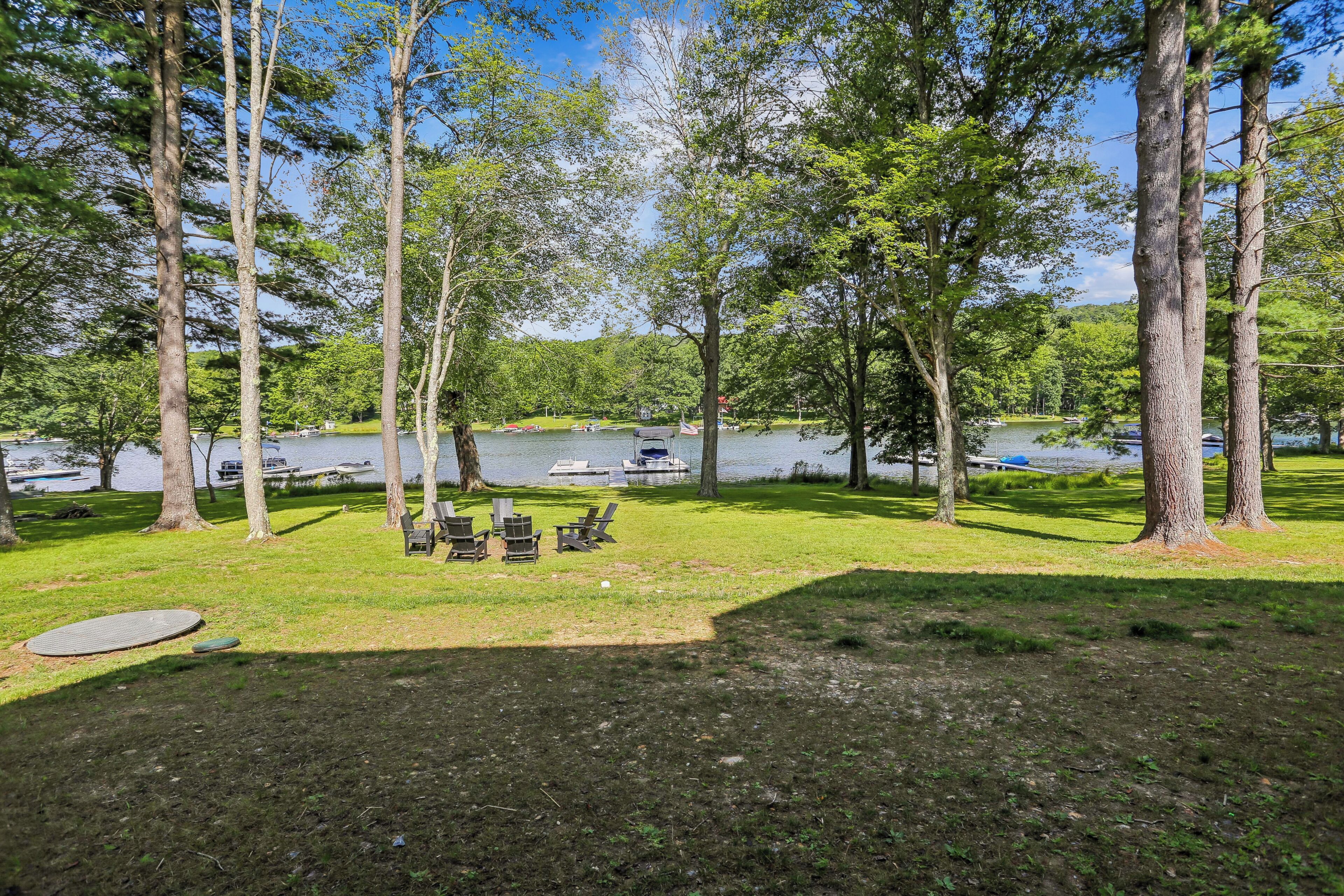Ein Campingplatz am Seeufer mit Picknicktischen und -stühlen, umgeben von hohen Bäumen und Booten auf dem Wasser.