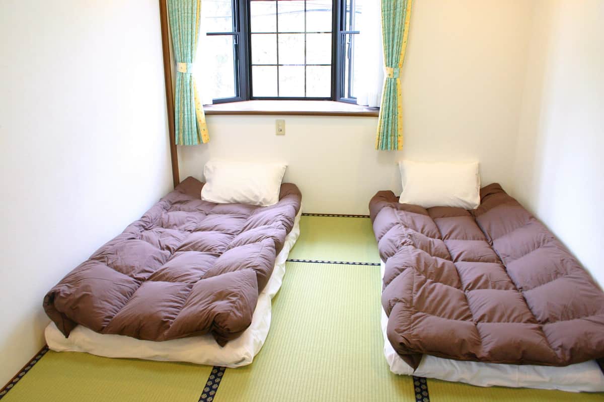 Quarto com duas camas de solteiro, cada uma com travesseiro e cobertor. Há uma janela com cortinas, e o chão é revestido com esteiras de tatame.