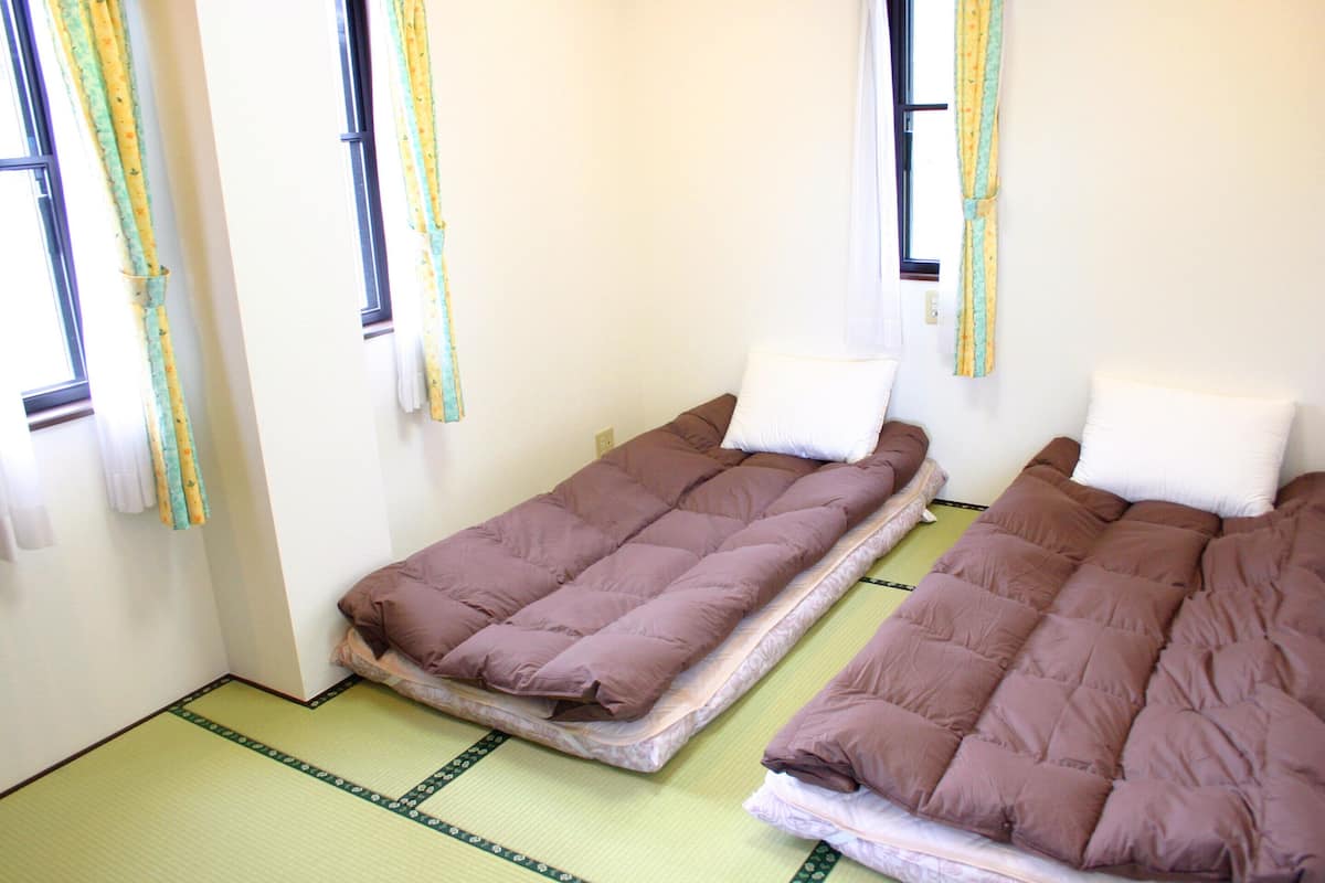 Duas camas de solteiro com almofadas, chão de esteira de tatame verde e janela com cortinas.