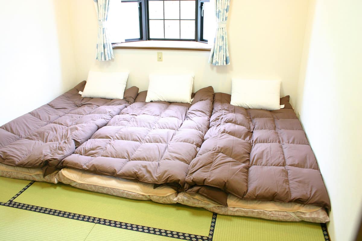 Uma cama com colcha marrom, janela com cortinas e piso de esteira de tatame.