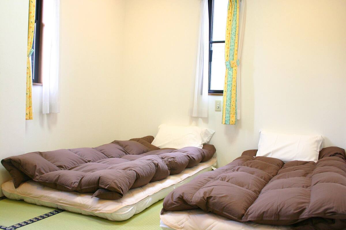 Duas camas de solteiro com roupa de cama marrom num quarto com paredes simples e janelas com cortinas.