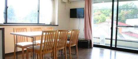 Un comedor con una mesa y sillas de madera, un televisor de pantalla plana en la pared y un ventanal con vistas a árboles y edificios.