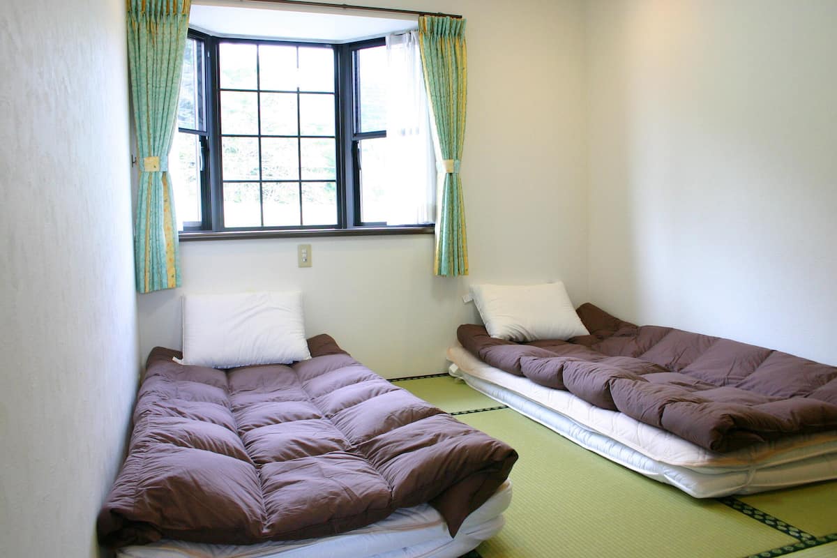 Duas camas de solteiro com colchões, uma janela com cortinas e uma tomada na parede.