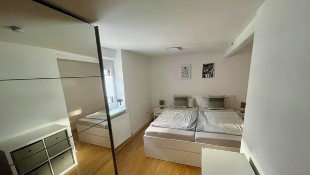 A modern bedroom with a large bed, a small bedside table, a radiator, and a window. - Vacation apartment at Walberla (Kirchehrenbach)