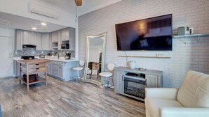 Une cuisine moderne avec des meubles blancs, un îlot central en bois et un grand téléviseur à écran plat fixé à un mur en briques.