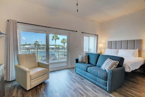 A hotel room with a bed, a sofa, a chair, and a large window with a view of palm trees and a balcony.