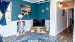 Ein Wohnzimmer mit türkisfarbenen Wänden, einem Flachbildfernseher, einem Bücherregal und einem blauen Teppich mit Muster.