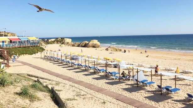 Una playa con tumbonas y sombrillas, un muelle de madera y una costa rocosa.