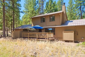 A two-story wooden cabin with a deck, surrounded by tall pine trees. - 8 East Butte - Heart of Sunriver, Game Room, Hot Tub, Bikes, SHARC, A/C (Bend)