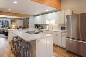 A modern kitchen with a central island, bar stools, a stainless steel refrigerator, and wooden flooring. - 8 East Butte - Heart of Sunriver, Game Room, Hot Tub, Bikes, SHARC, A/C (Bend)