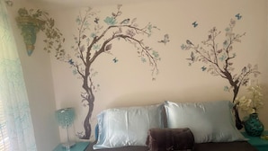 Ein Schlafzimmer mit einem Bett, zwei Kissen, einem Nachttisch, einer Lampe und einer Wandmalerei mit Bäumen und Vögeln.