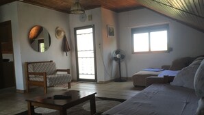 Ein gemütliches Zimmer mit Holzbalkendecke, einem runden Spiegel, einem gestreiften Sessel, einem Bett, einem Tisch und einem Standventilator.