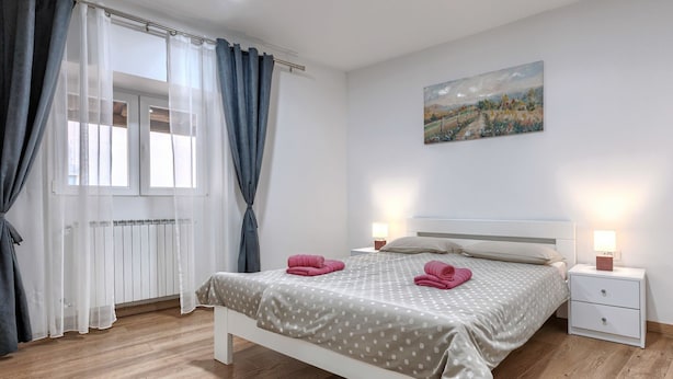 A bedroom with a bed, two bedside tables, a window with curtains, a radiator, and a painting on the wall.