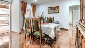 A dining room with a table set for four, surrounded by wooden chairs with green upholstery. A painting hangs on the wall above the table, and a wooden cabinet is to the right.