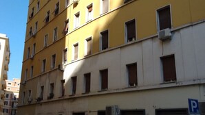 Ein mehrgeschossiges Gebäude mit mehreren Fenstern, von denen einige geöffnet sind. Klimaanlagen sind an der Fassade angebracht, und ein Parkschild ist sichtbar.