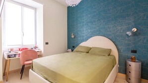 Ein Schlafzimmer mit einem Bett, einem Schreibtisch, einem Sessel und einem Nachttisch, an der Wand ist ein Blattmuster zu sehen.
