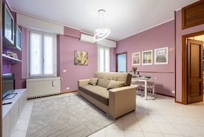 Un salon comprenant un canapé, une table à manger et des chaises. On y trouve également un radiateur, une fenêtre avec des rideaux et un luminaire au plafond.