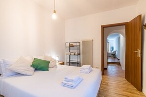 A hotel room with a bed, a bedside table, a lamp, a radiator, a shelf with towels, and a wooden door.