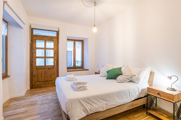 Une chambre à coucher avec un lit en bois, une table de chevet, une lampe de bureau et deux fenêtres.