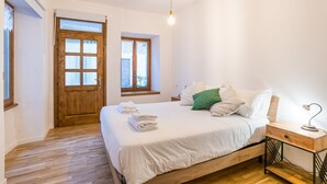 Quarto com cama de madeira, mesa de cabeceira, luminária de escrivaninha e duas janelas.