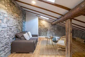 A cozy attic room with exposed wooden beams, a stone wall, a brown sofa, a wicker chair, and a small round table.