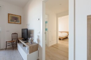 Une pièce comprenant un bureau en bois, une chaise, un téléviseur et un vase. La pièce adjacente contient un lit avec un matelas et des oreillers blancs.