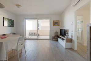 A spacious living area with a dining table, chairs, a TV, and a sliding glass door leading to a balcony.
