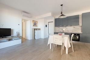 A modern living room with a flat-screen TV, a white dining table with chairs, a kitchen with grey cabinets and a marble backsplash, and a hallway with a door.