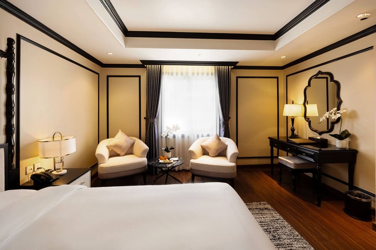 A hotel room with a large bed, two armchairs, a small table with a lamp, a desk with a mirror, and a window with curtains.