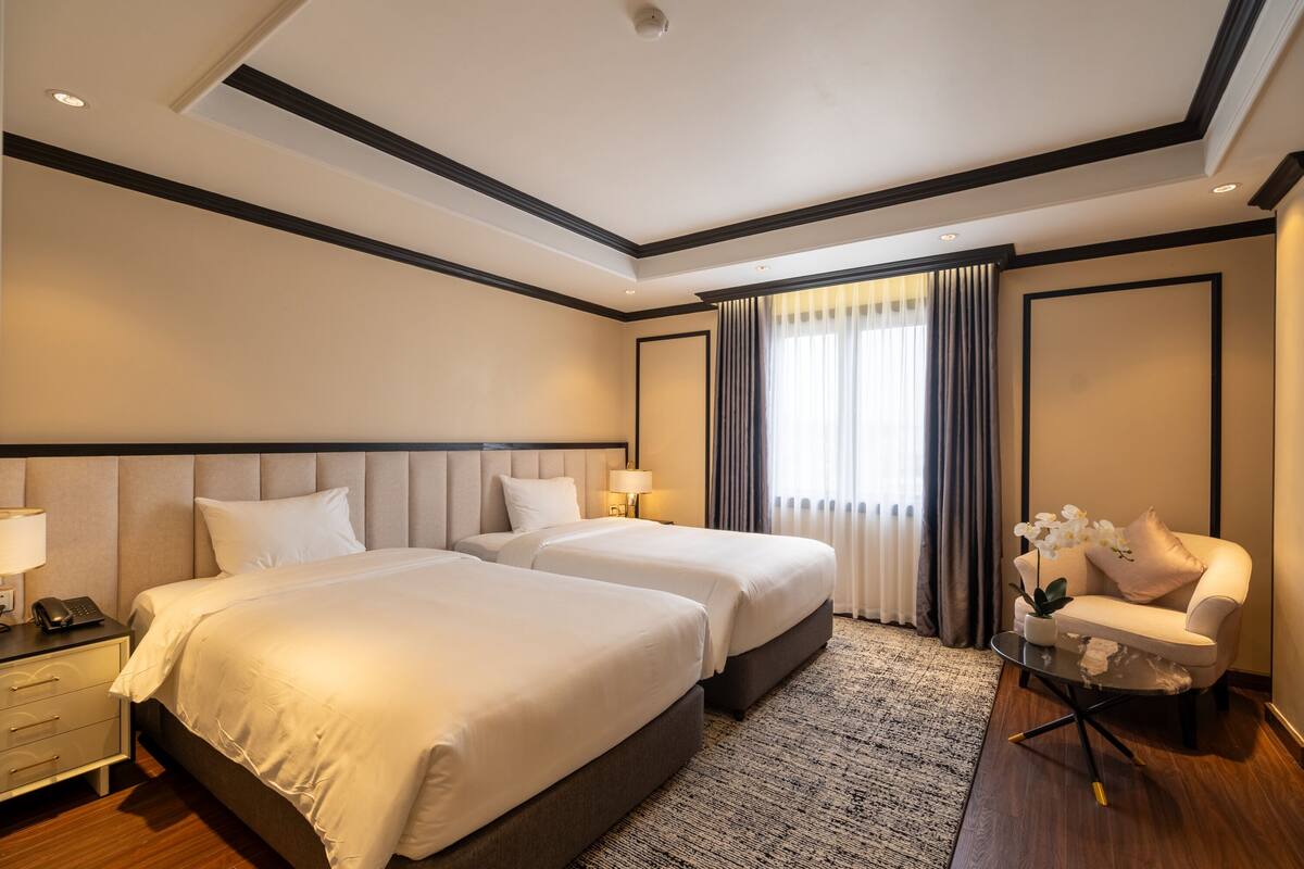 A hotel room with two beds, a nightstand with a lamp, a small table with a plant, and a chair.