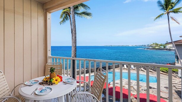 A balcony with a table set for dining, overlooking a pool and the ocean. - Sea Village Oceanview Condo (Kailua-Kona)