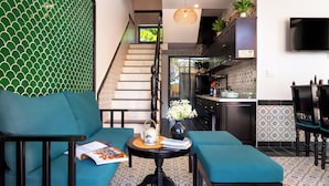 Una sala de estar moderna con sofás color turquesa, una pequeña mesa redonda con un florero y una revista, una escalera con papel tapiz estampado, y una zona de cocina con televisión y señal de WiFi.